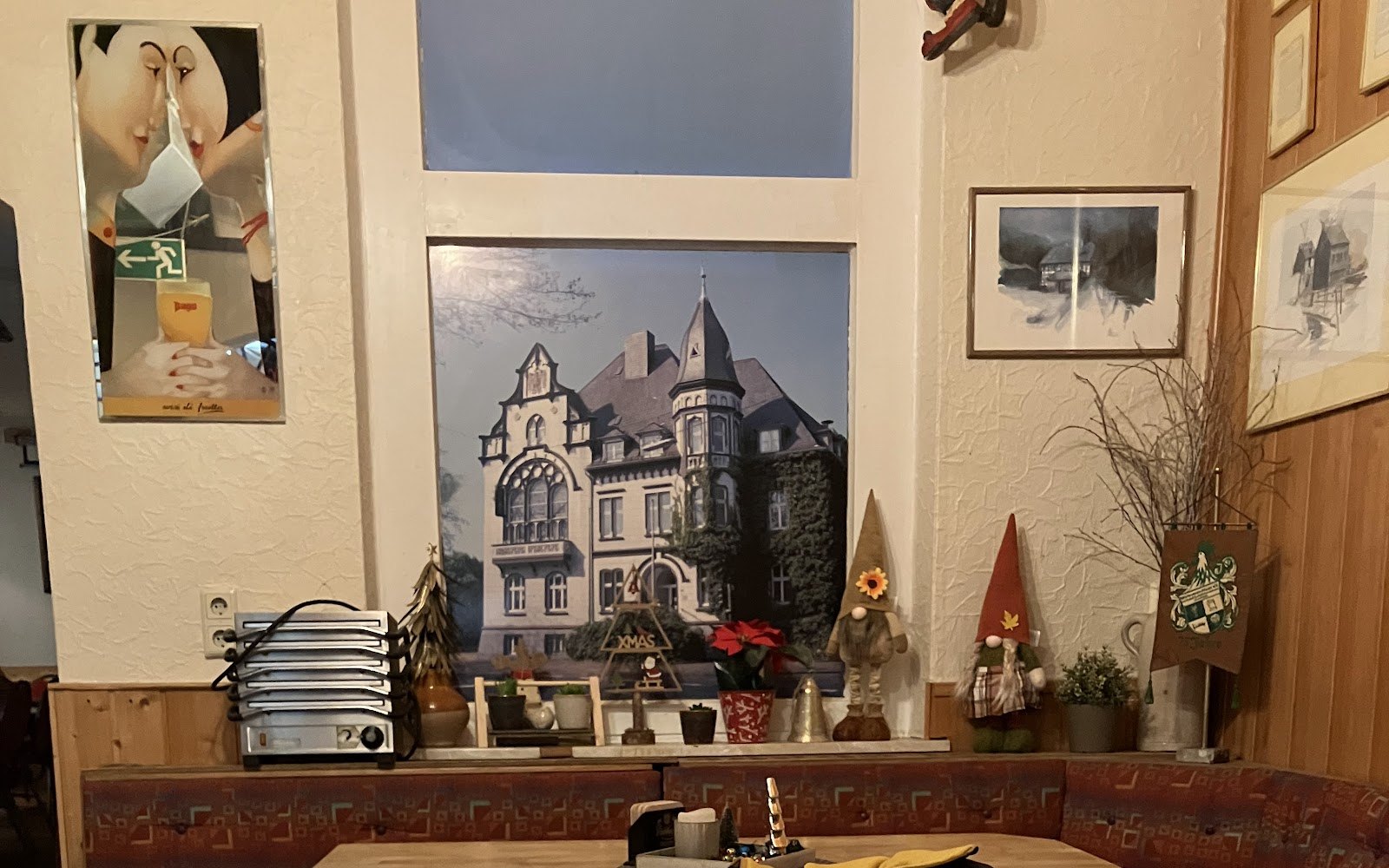 Die Gaststube mit gepolsterter Eckbank, einem großen Bild einer Villa an der Wand und Wichtelfiguren auf der Fensterbank