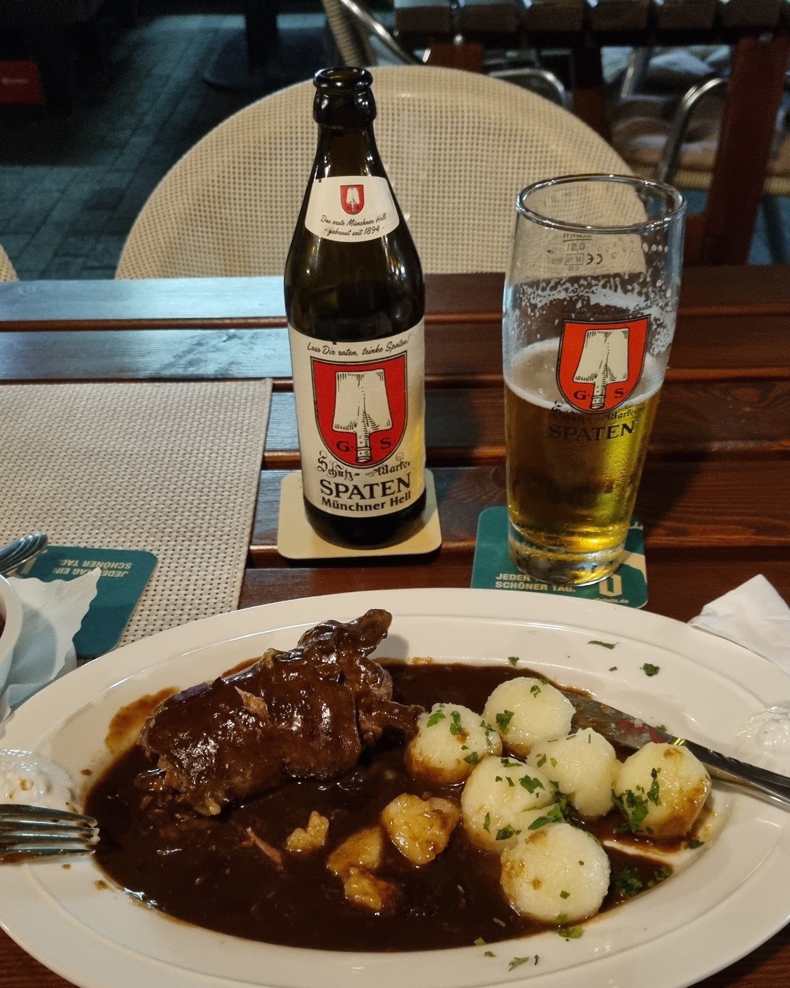 Braten in dunkler Soße mit kleinen Kartoffeln, dahinter ein frisch gezapftes Bier