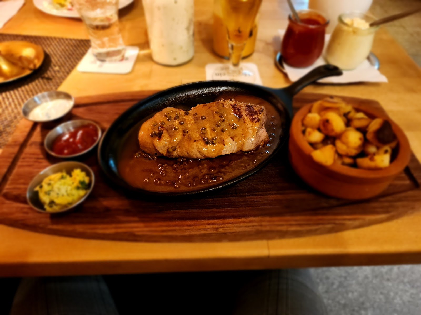 Steak mit Soße in der Pfanne, Bratkartoffeln im Tontopf und Dips auf dem Holzbrett